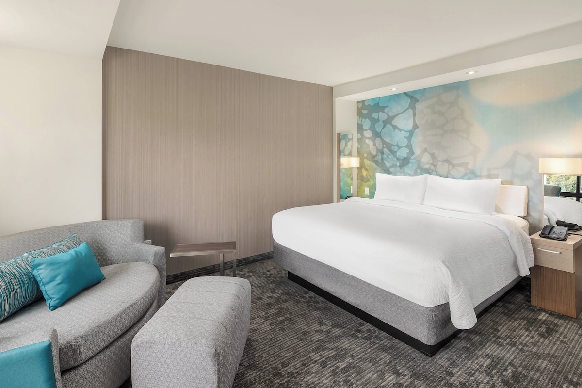 A hotel room with a large bed, a sofa, a small table, and a nightstand with a lamp.