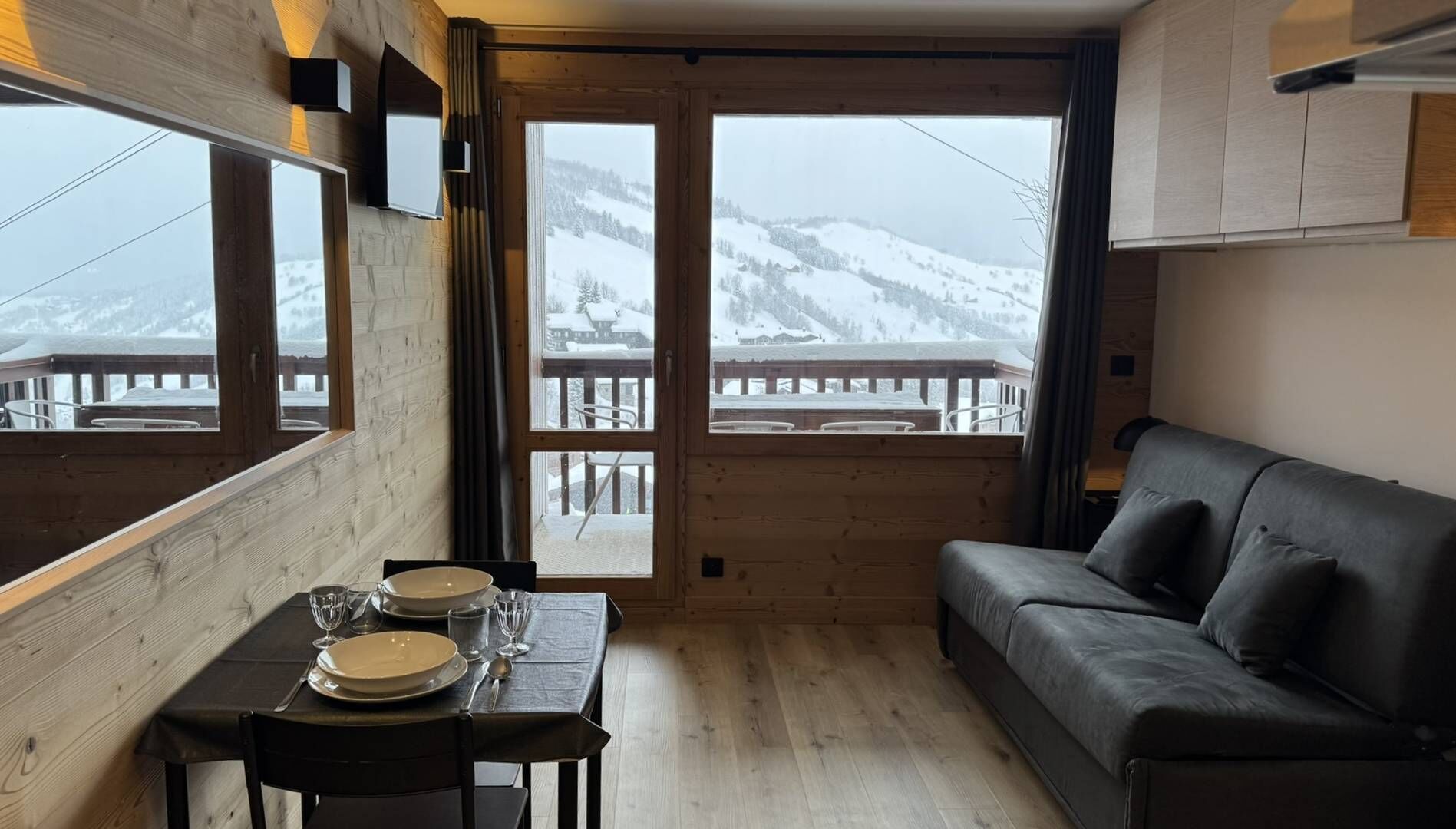 Interior acogedor de una cabaña con una mesa de comedor preparada para dos personas, un sofá y vistas a montañas nevadas a través de amplios ventanales.
