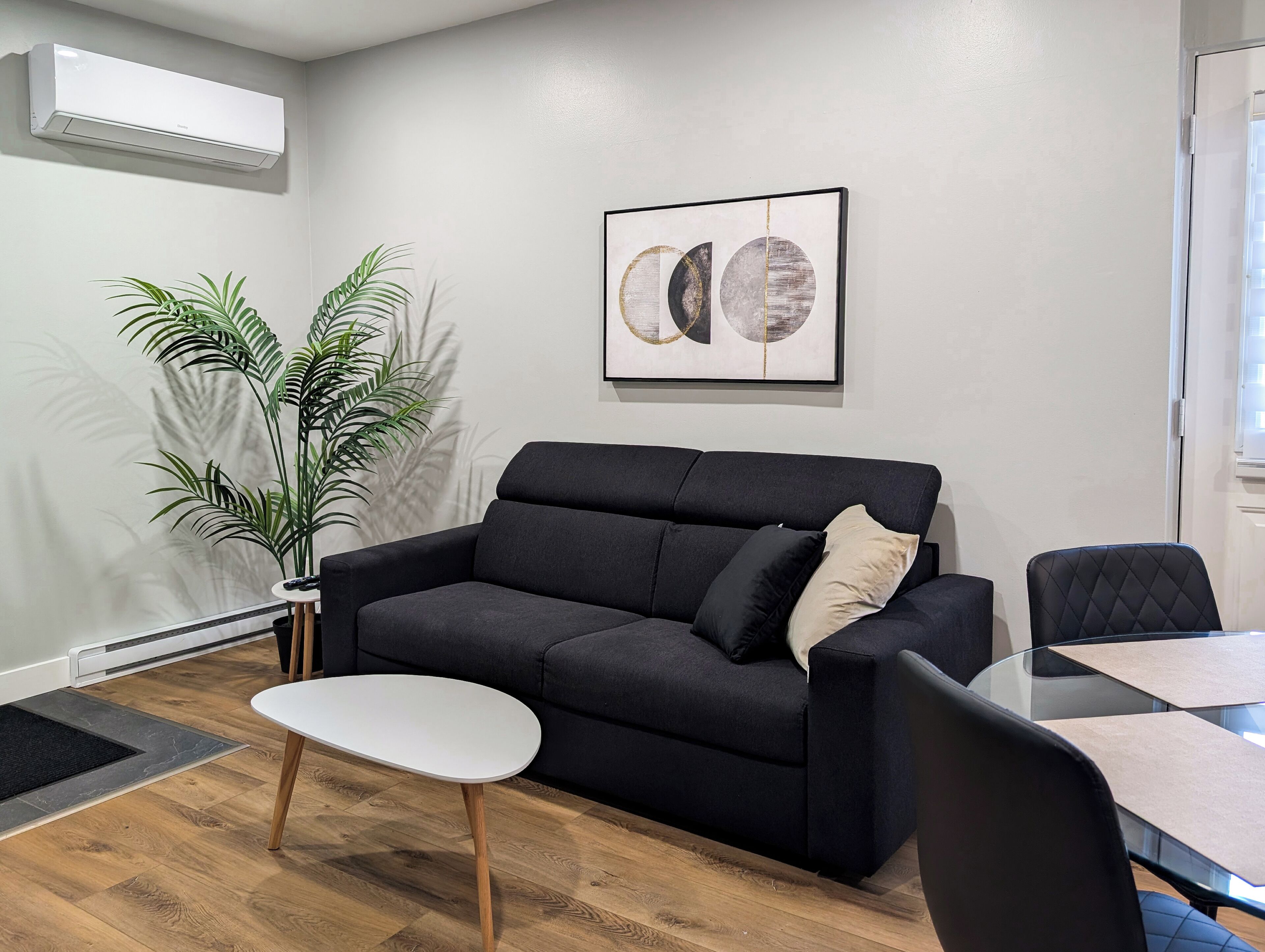 A modern living room with a black sofa, a white coffee table, and a glass dining table. There is a large potted plant and a framed abstract artwork on the wall.