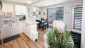 A modern kitchen with white cabinets, a dining area with a table and chairs, and a large window with blinds. - Hébergement Harvey (Alma)