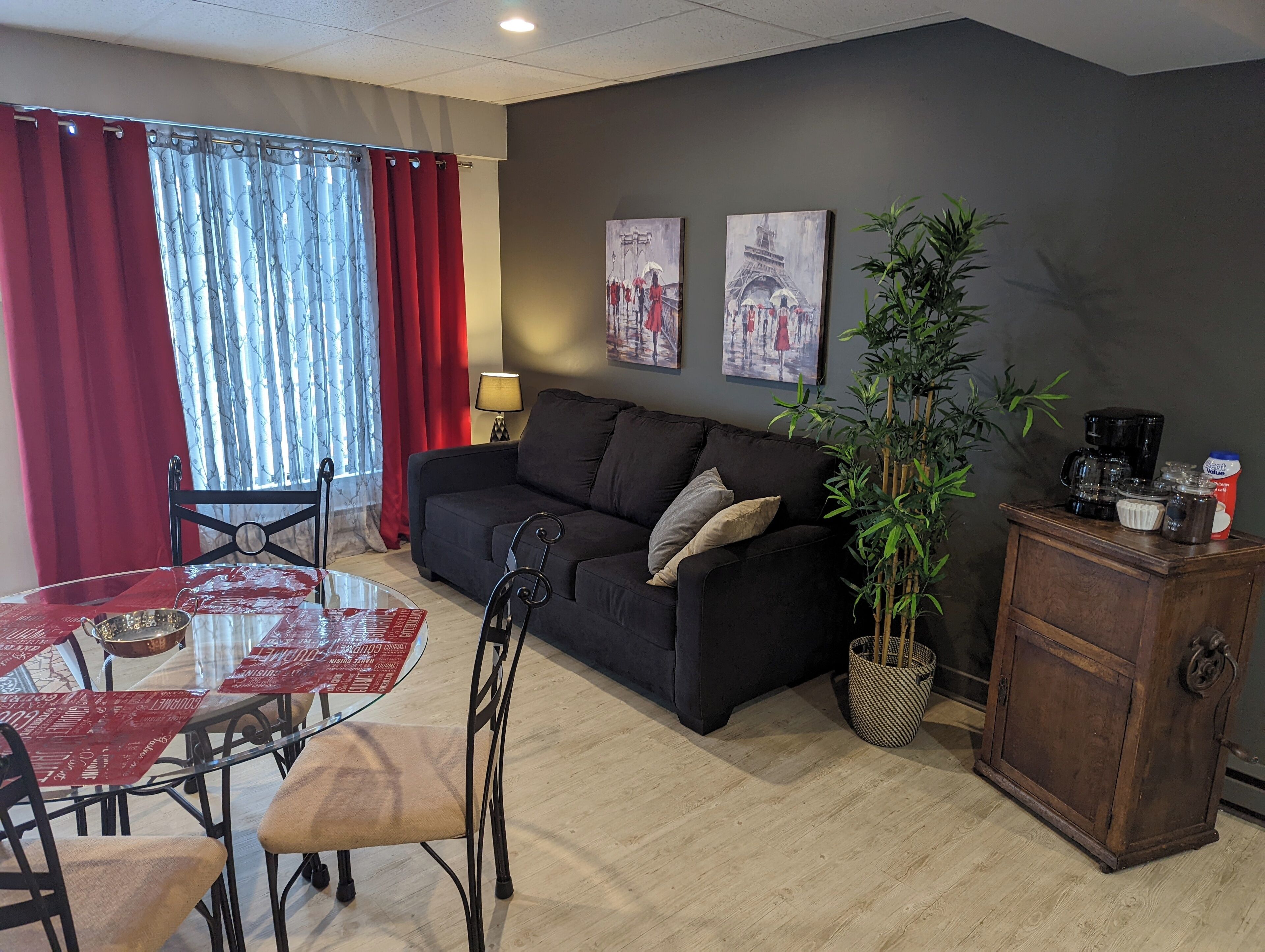 A living room with a black sofa, a glass coffee table, and a dining table with chairs. There are two paintings on the wall, a lamp, and a bamboo plant.