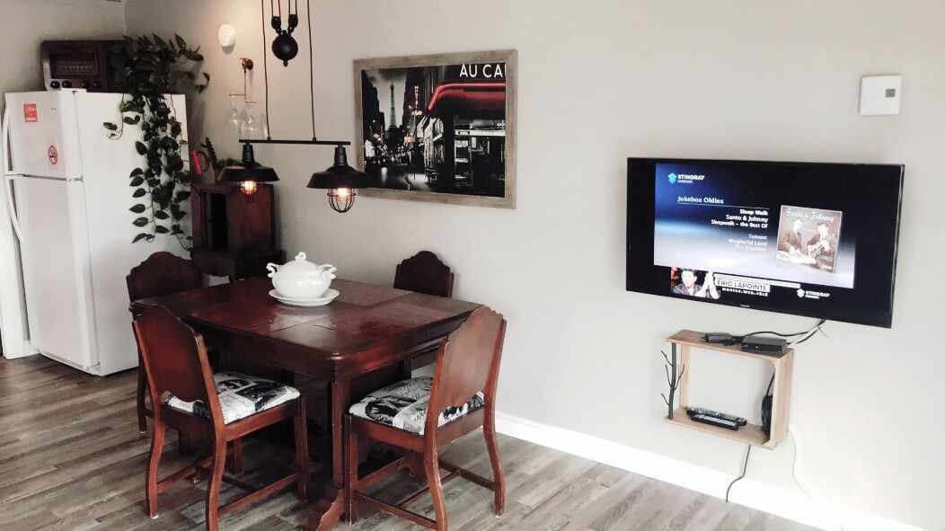 A dining area with a wooden table, chairs, a white refrigerator, a TV mounted on the wall, and a small shelf with electronic devices.