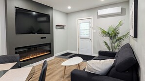 A modern living room with a flat-screen TV, a fireplace, a dining area with a glass table, and a large potted plant. - Hébergement Harvey (Alma)
