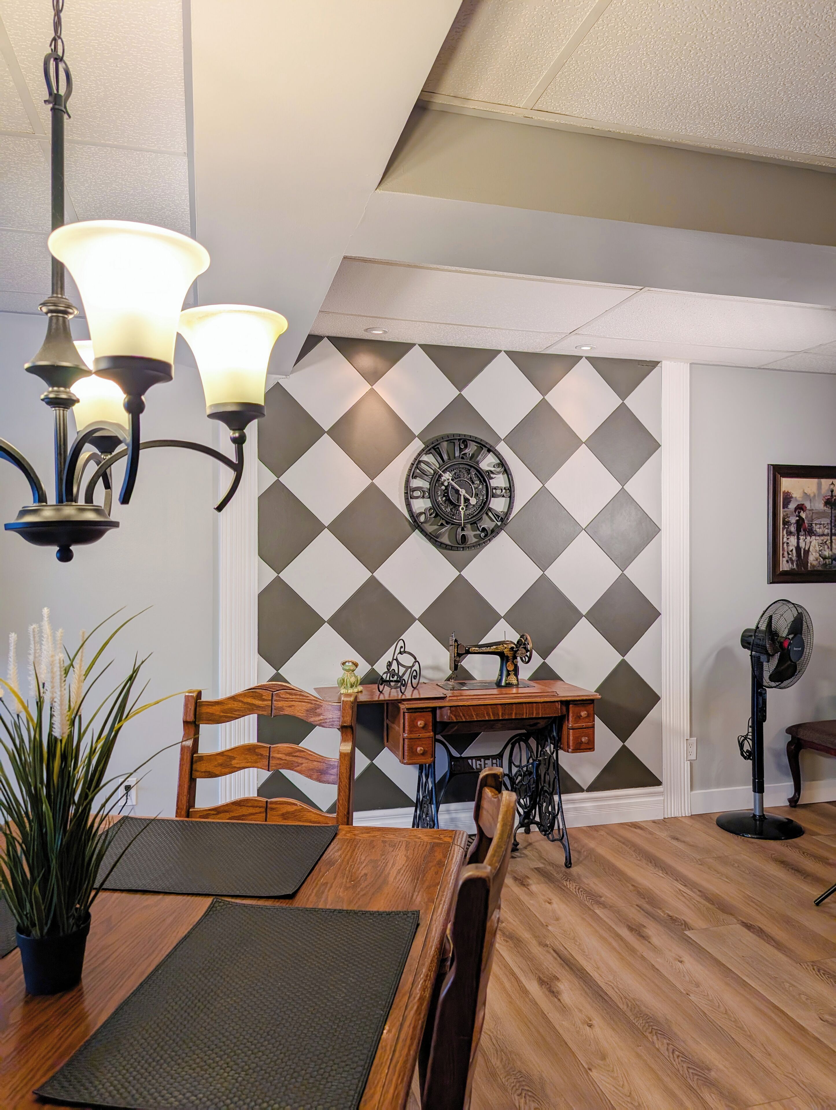 A dining area with a wooden table, chairs, a chandelier, a wall clock, and a sewing machine.