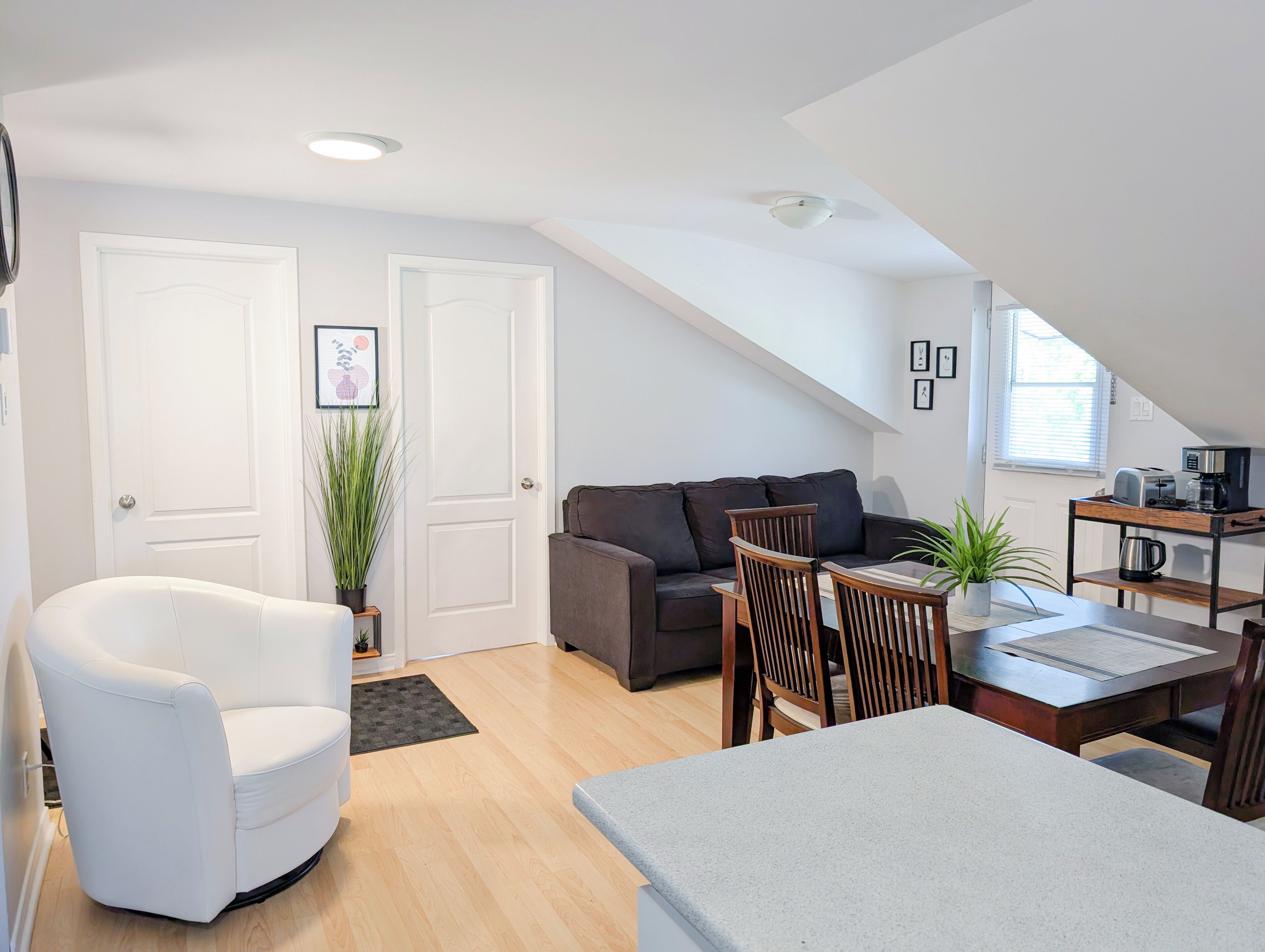 Una sala moderna con un sofá, un sillón blanco, una mesa de comedor con sillas, una cafetera y una pequeña cocina con un tostador y hervidor.