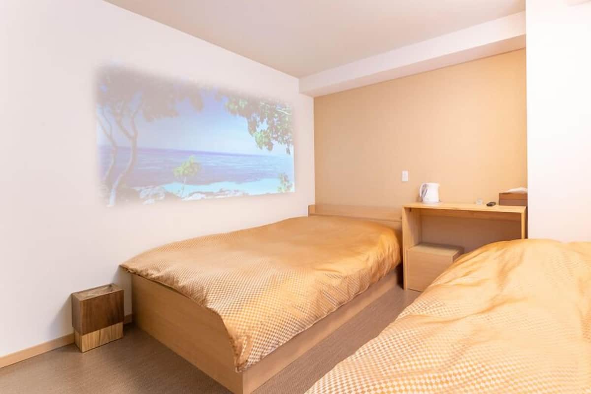 Una habitación con dos camas, un escritorio pequeño, un proyector en la pared que muestra una escena de playa y una mesita de noche de madera.