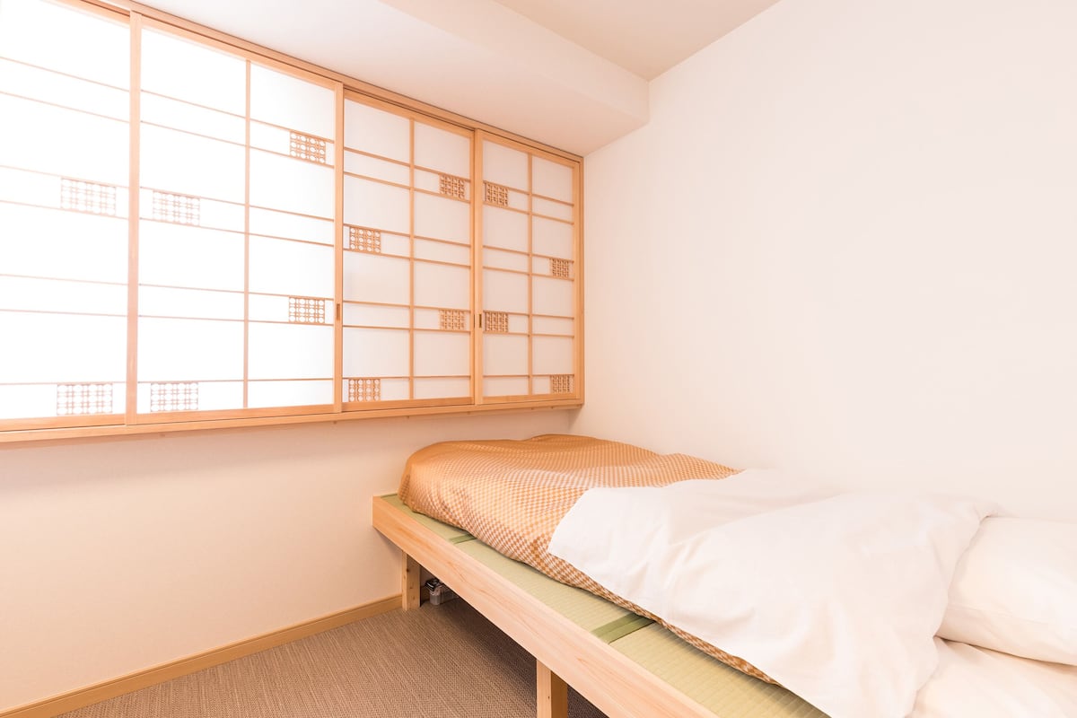 Una habitación japonesa tradicional con suelo de tatami, una cama baja de madera con colchón a cuadros y una puerta corredera tipo shoji.