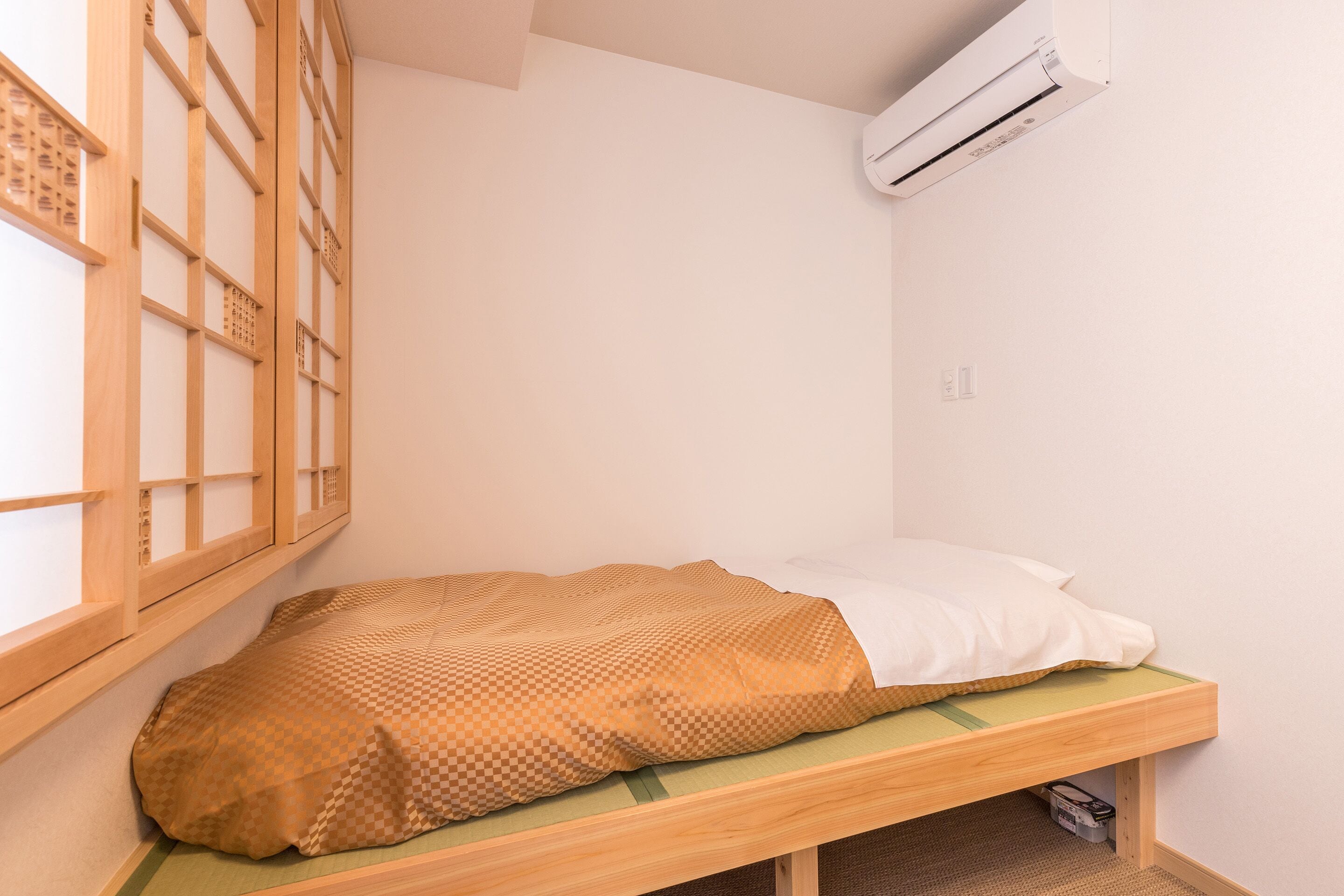 A small, single-bed room with a wooden bed frame, a checkered blanket, and a white pillow. An air conditioner is mounted on the wall above the bed.