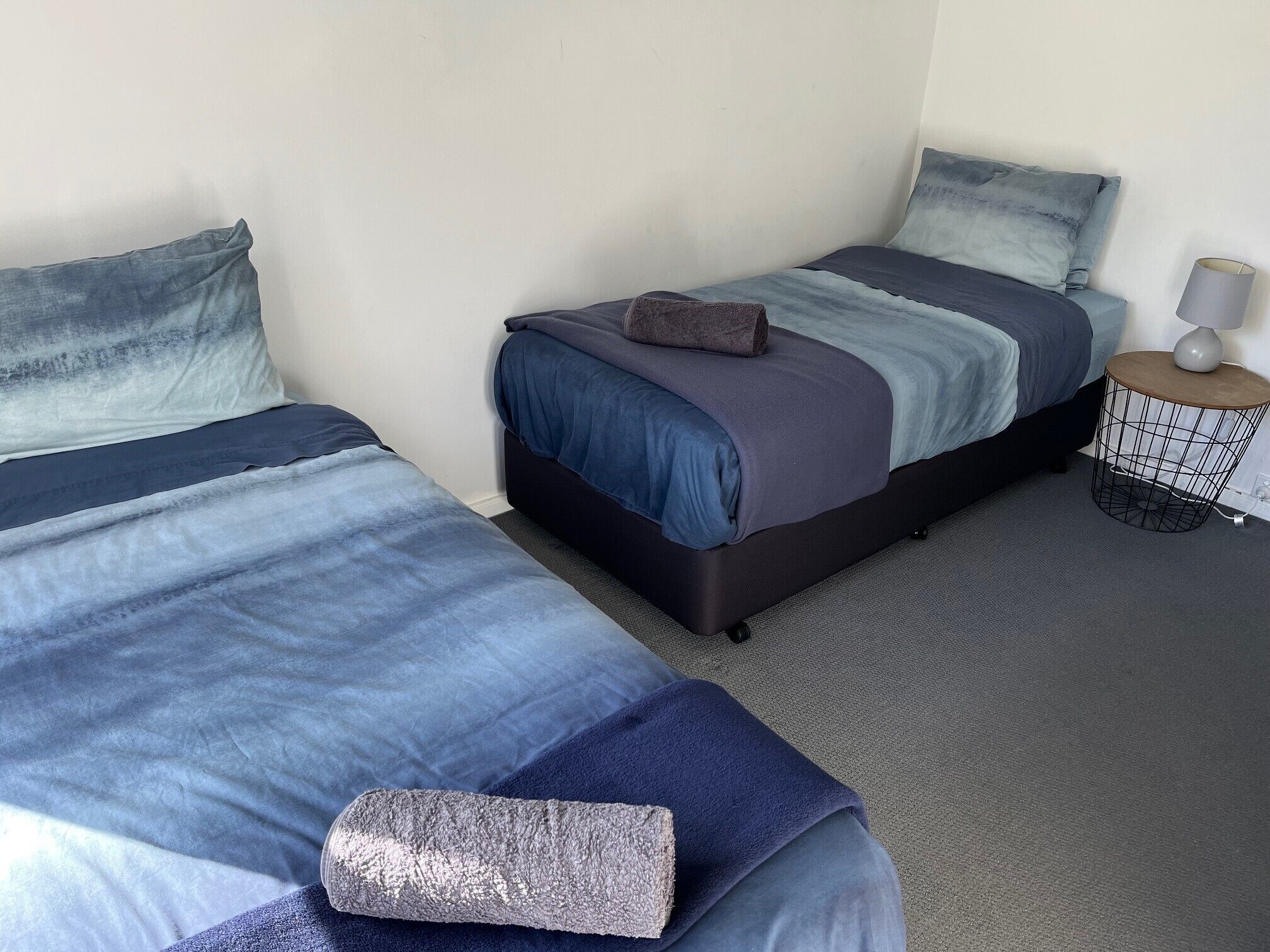 Hotelkamer met twee bedden, elk met een blauw-grijze overtrek, een klein rond nachtkastje met een lamp, en een grijze vloerbedekking.
