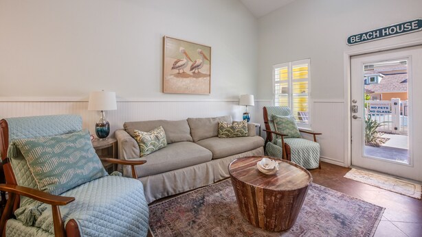 A living room with a sofa, two armchairs, a coffee table, a painting, and a door labeled 'BEACH HOUSE'.