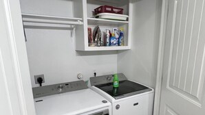 Une petite buanderie équipée d’un lave-linge, d’un sèche-linge et d’une étagère située au-dessus, où sont rangés les produits de nettoyage.