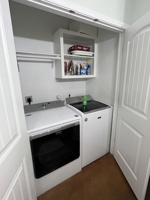 A compact laundry room with a washing machine, dryer, and a shelf above containing cleaning supplies.