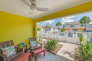 A yellow room with two wooden chairs and a bench, a ceiling fan, and a view of a fenced area with houses and palm trees.