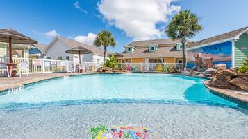 A clear swimming pool with a tropical setting, surrounded by colorful buildings and a white fence.