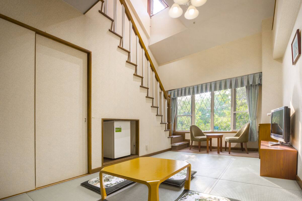 A room with tatami flooring, a low wooden table, a television, a refrigerator, and a staircase leading to an upper floor.