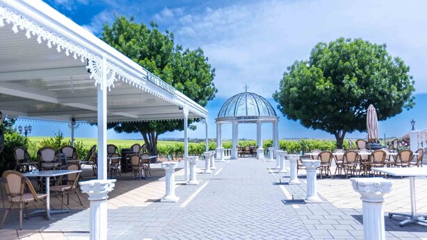 Een terras buiten met witte zuilen, tafels en stoelen, dat leidt naar een gazebo met een koepeldak.