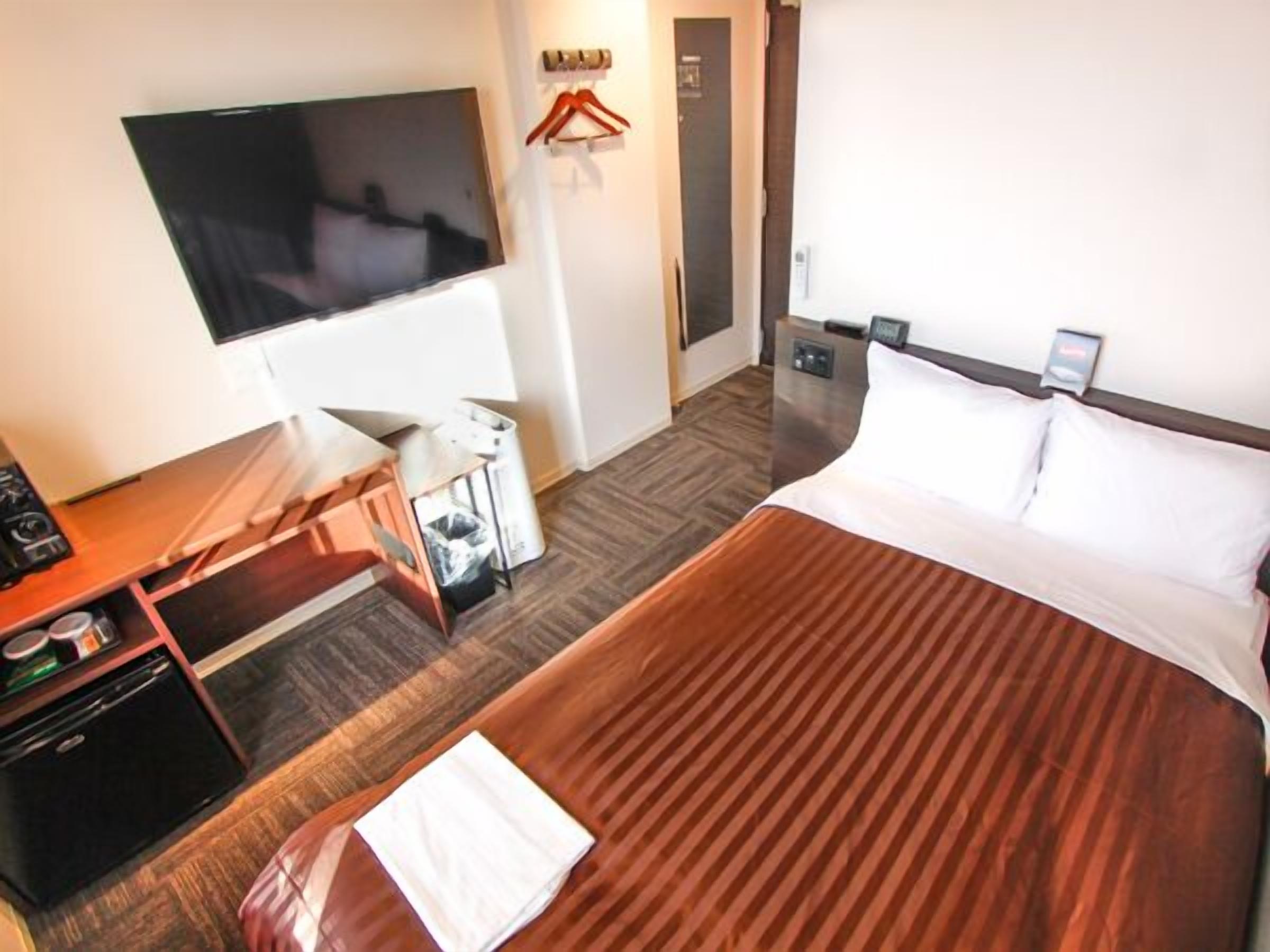 Habitación de hotel con cama, televisor montado en la pared, escritorio con silla y un pequeño cubo de basura.