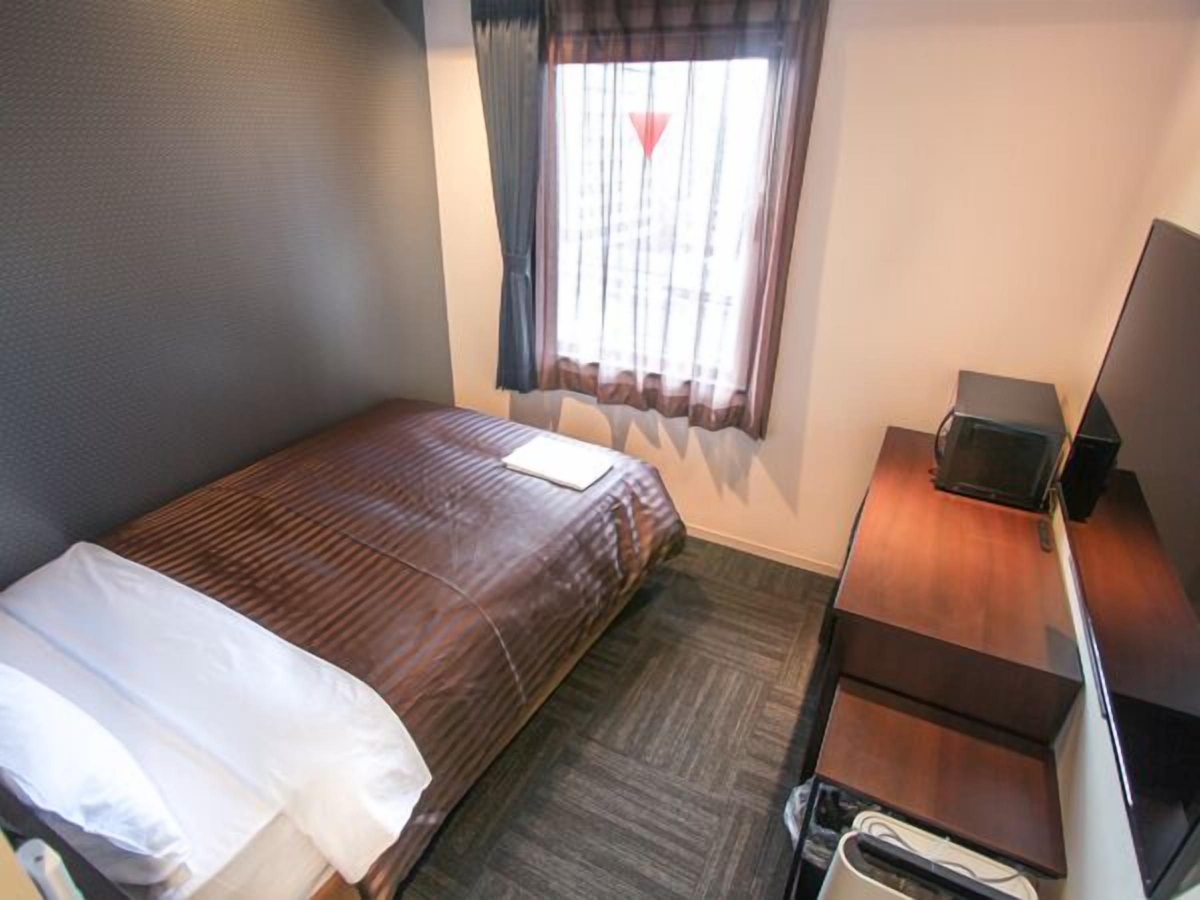 Una habitación de hotel pequeña y ordenada con una cama individual, un escritorio con computadora y una ventana con cortinas.