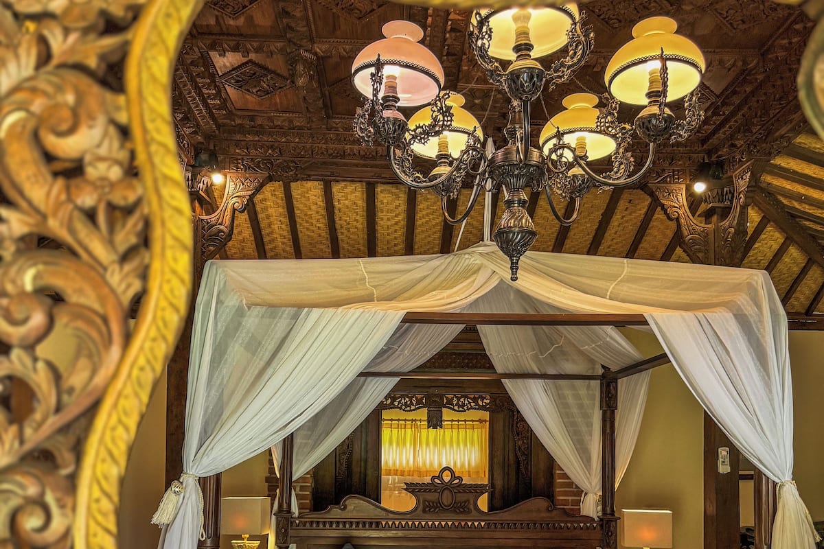 Ein Himmelsbett mit aufwendigem Holzkopf- und Fußteil, einer Chandelierleuchtre und einem dekorativen Spiegel über dem Bett.