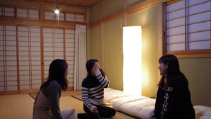Tres personas sentadas sobre esteras de tatami en una habitación con paneles de shoji y un gran farol de papel iluminado.