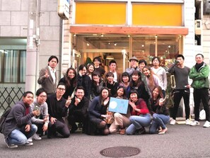 A group of people posing in front of a building with a glass entrance and a yellow awning. - Ini Kobe Hostel & Cafe Bar (Kobe)