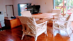 A room with a wooden dining table, white wicker chairs, a television, and a view of greenery outside. - Holiday House Sumire (Onna)