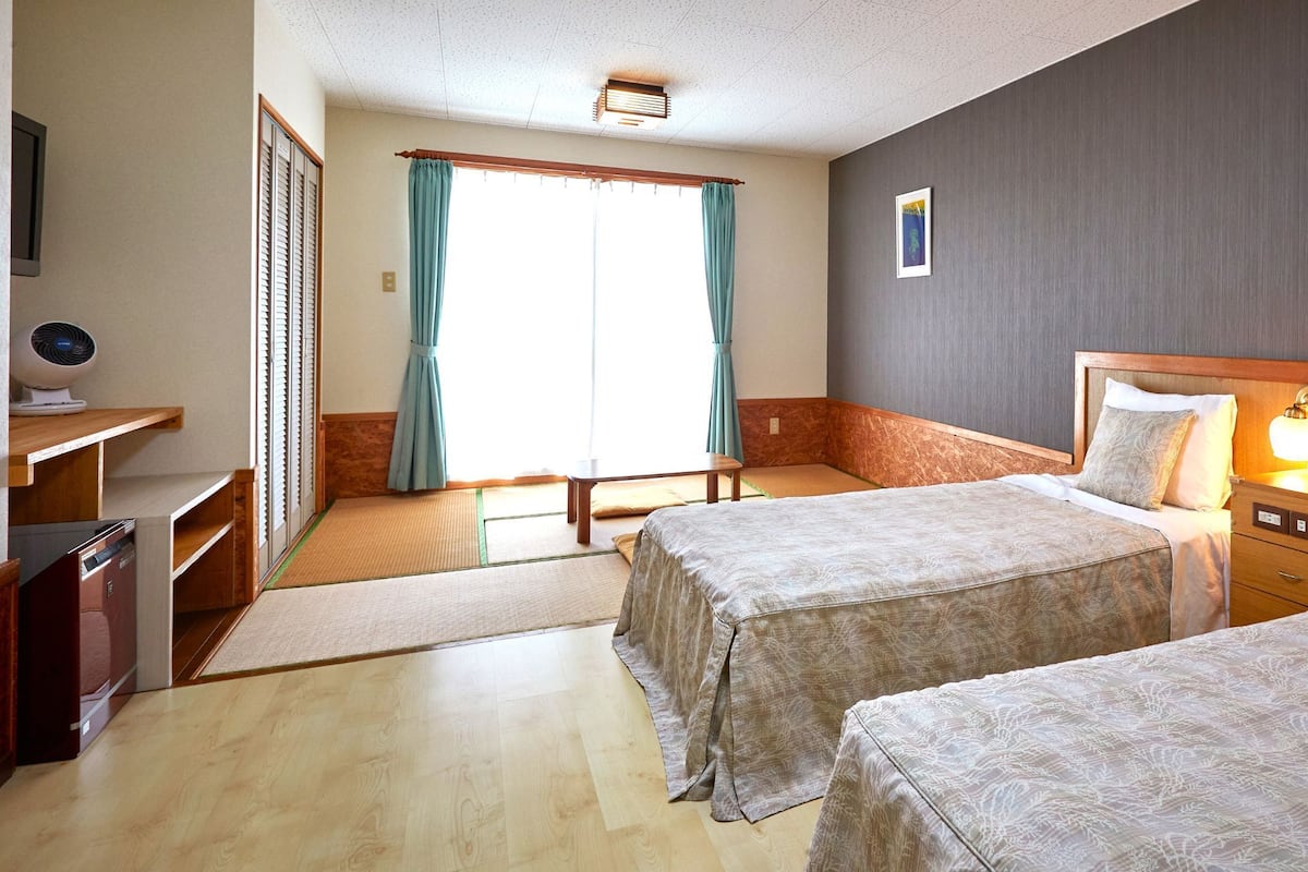 A hotel room with two beds, a desk, a chair, a small table, and a window with curtains.