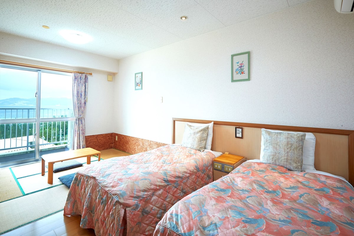 A hotel room with two beds, a bench, a view of the sea, and a balcony.