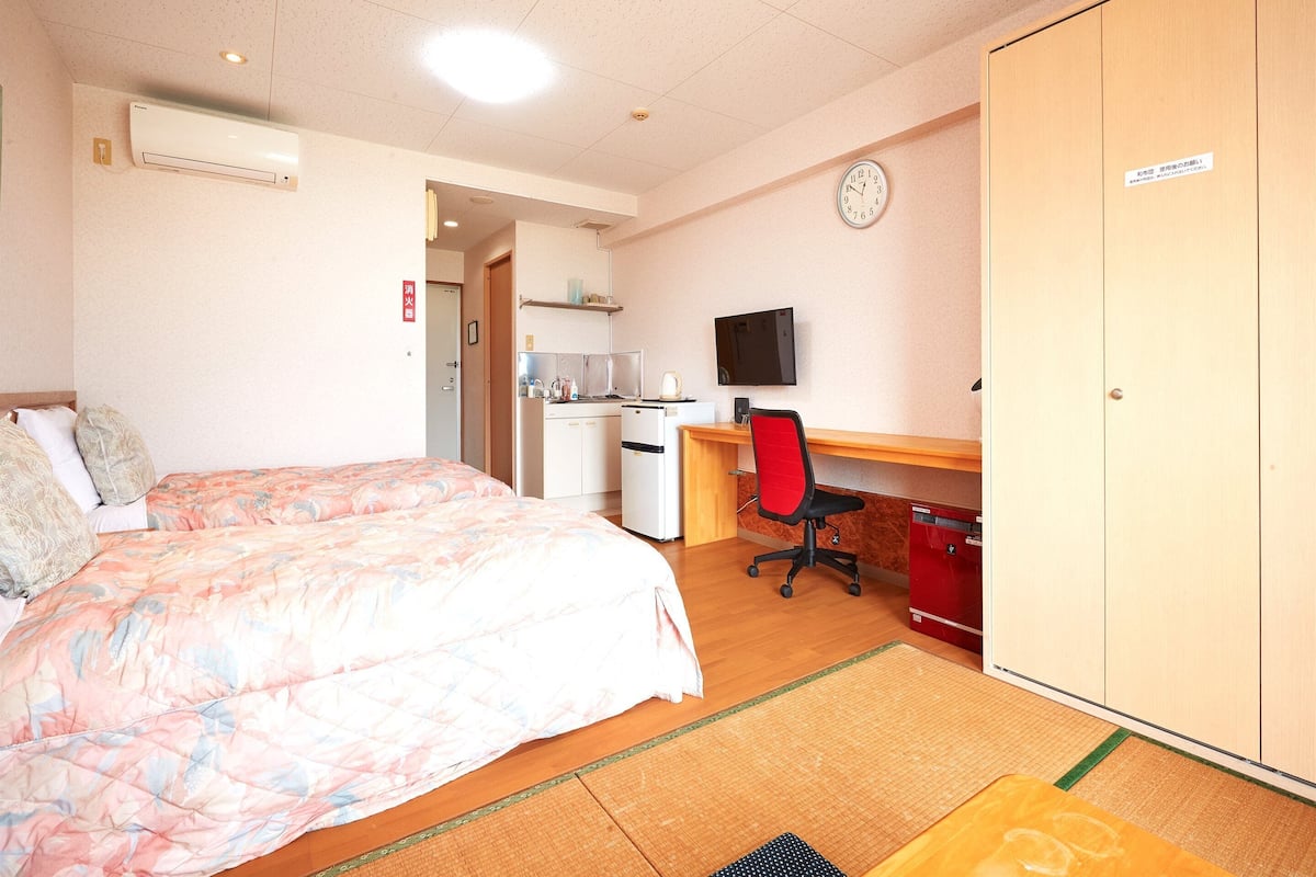 A hotel room with a bed, a desk with a chair, a television, a wardrobe, and a kitchenette.