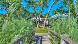 Un sentier en bois mène à une maison dotée d’un balcon, qui est entourée d’herbes hautes et d’arbres.