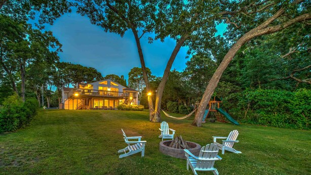Une maison avec une terrasse, un foyer extérieur et des fauteuils Adirondack dans le jardin.