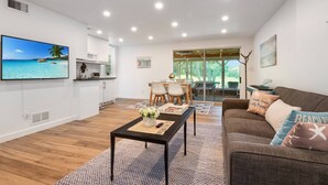 Un salon moderne avec un plancher en bois, une table basse noire, un canapé marron agrémenté de coussins aux motifs tropicaux, un téléviseur diffusant une scène de plage tropicale et un coin repas avec un piano.