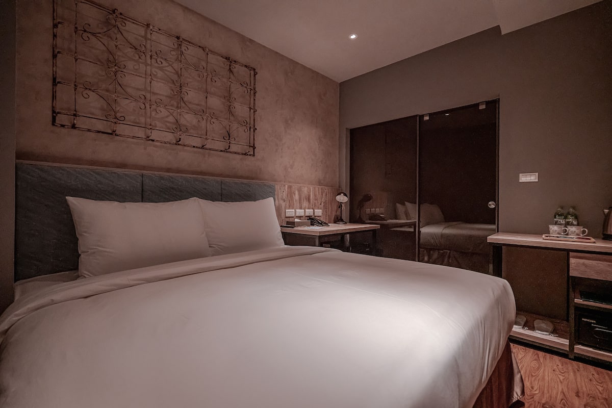 A modern hotel room with a large bed, a bedside table with a lamp, a mirror, and a wooden side table with a tray of cups.