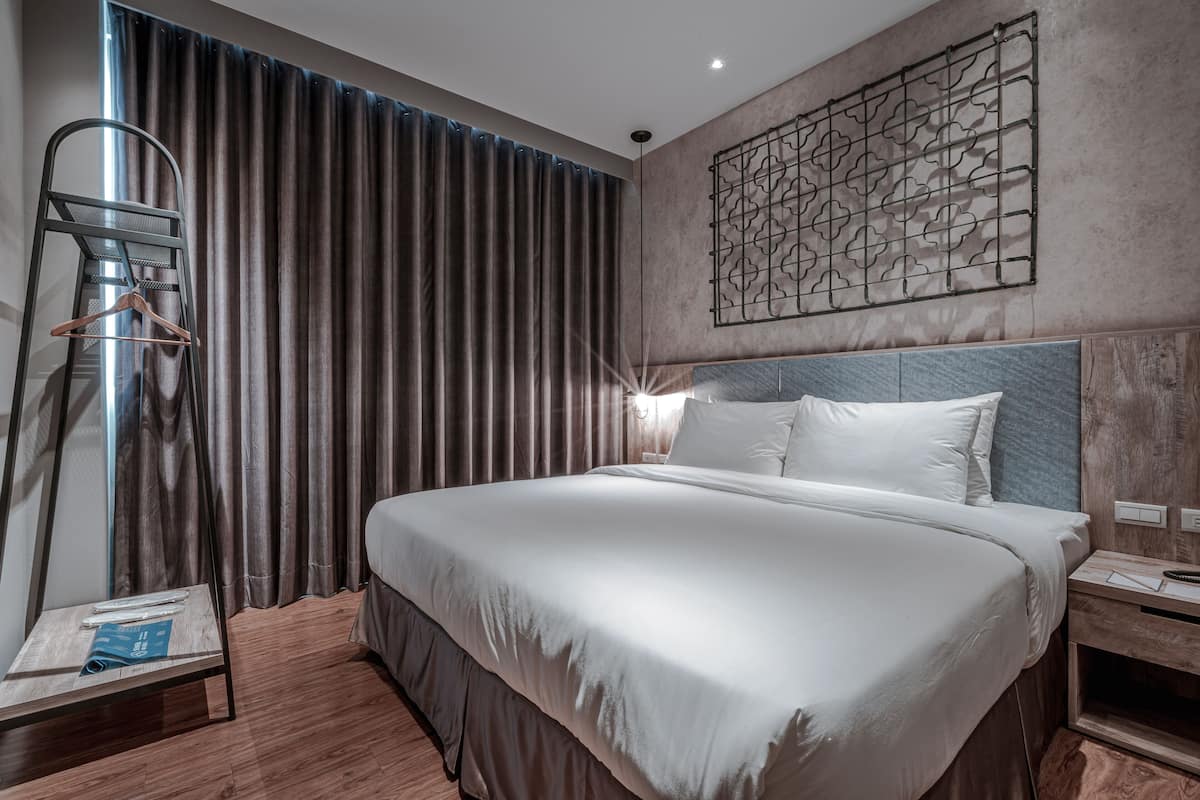 A modern hotel room with a large bed, a bedside table, a nightstand, a wall-mounted light fixture, and a decorative wall panel.