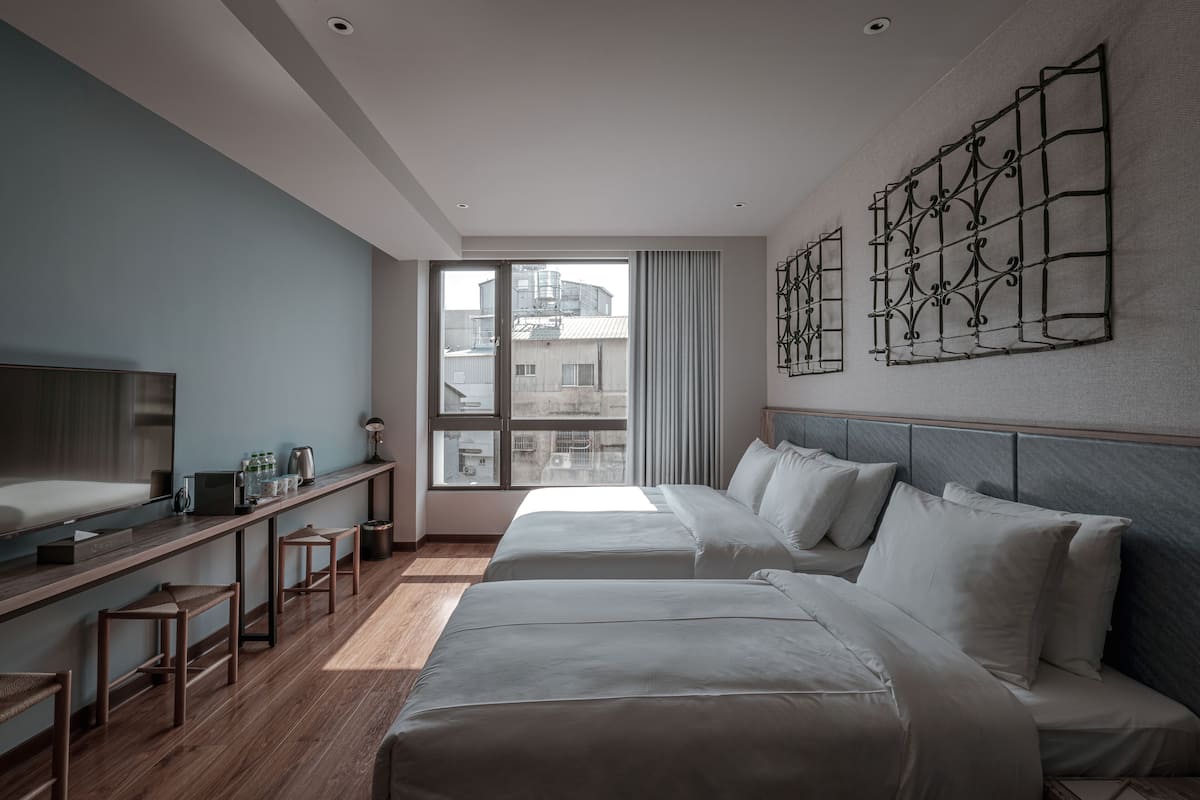 A modern hotel room with two beds, a large window, a TV, and a desk with stools.