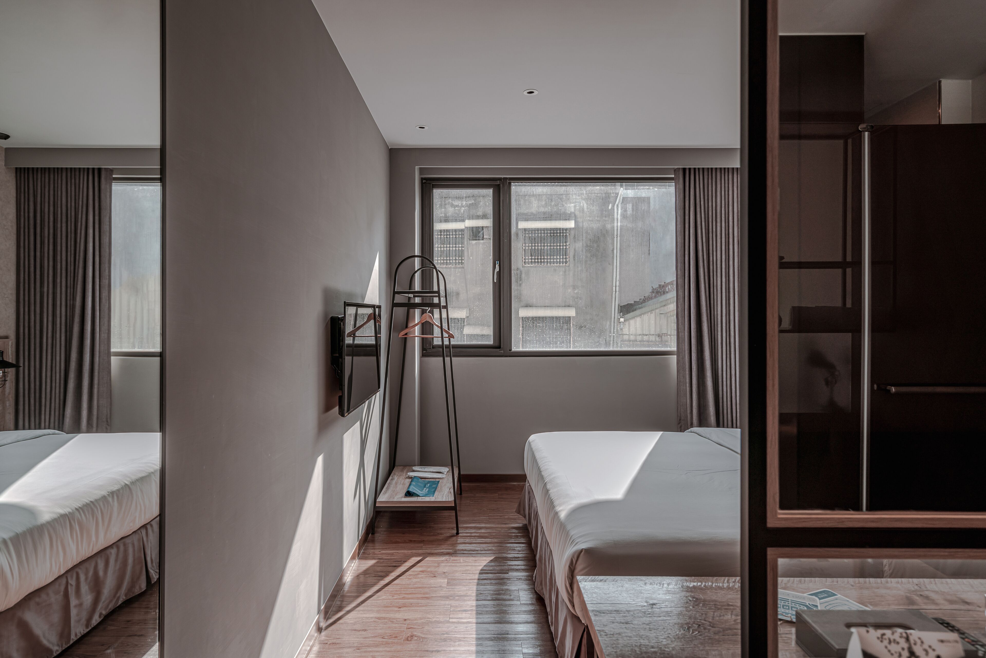 A modern hotel room with two single beds, a large window with a city view, a standing lamp, and a wardrobe.