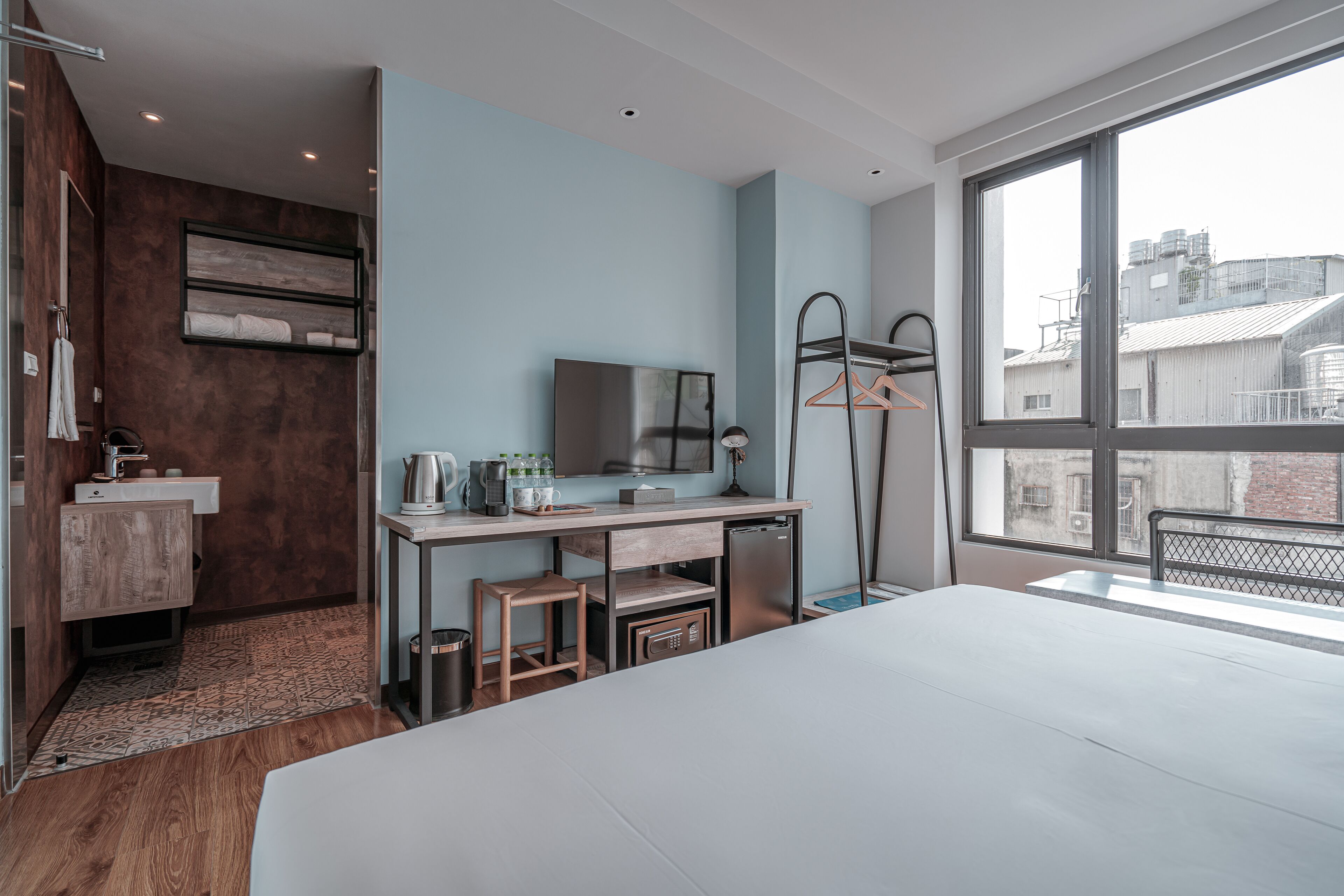 A modern hotel room with a large bed, a desk with a mirror, a small seating area, a wardrobe, and a view of the city through large windows.