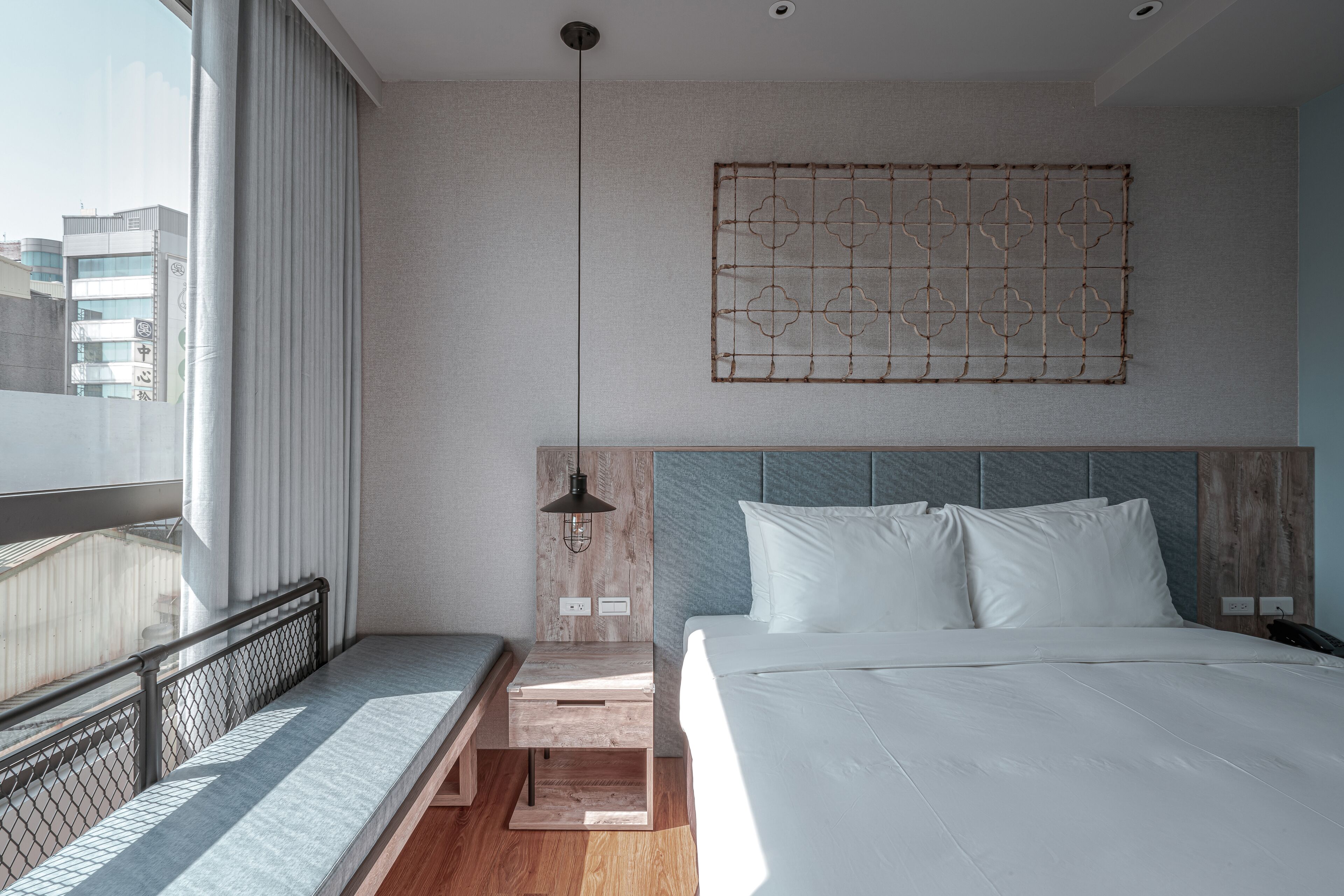 A modern hotel room with a large bed, a wooden bench, a view of the city, and a decorative wall piece.