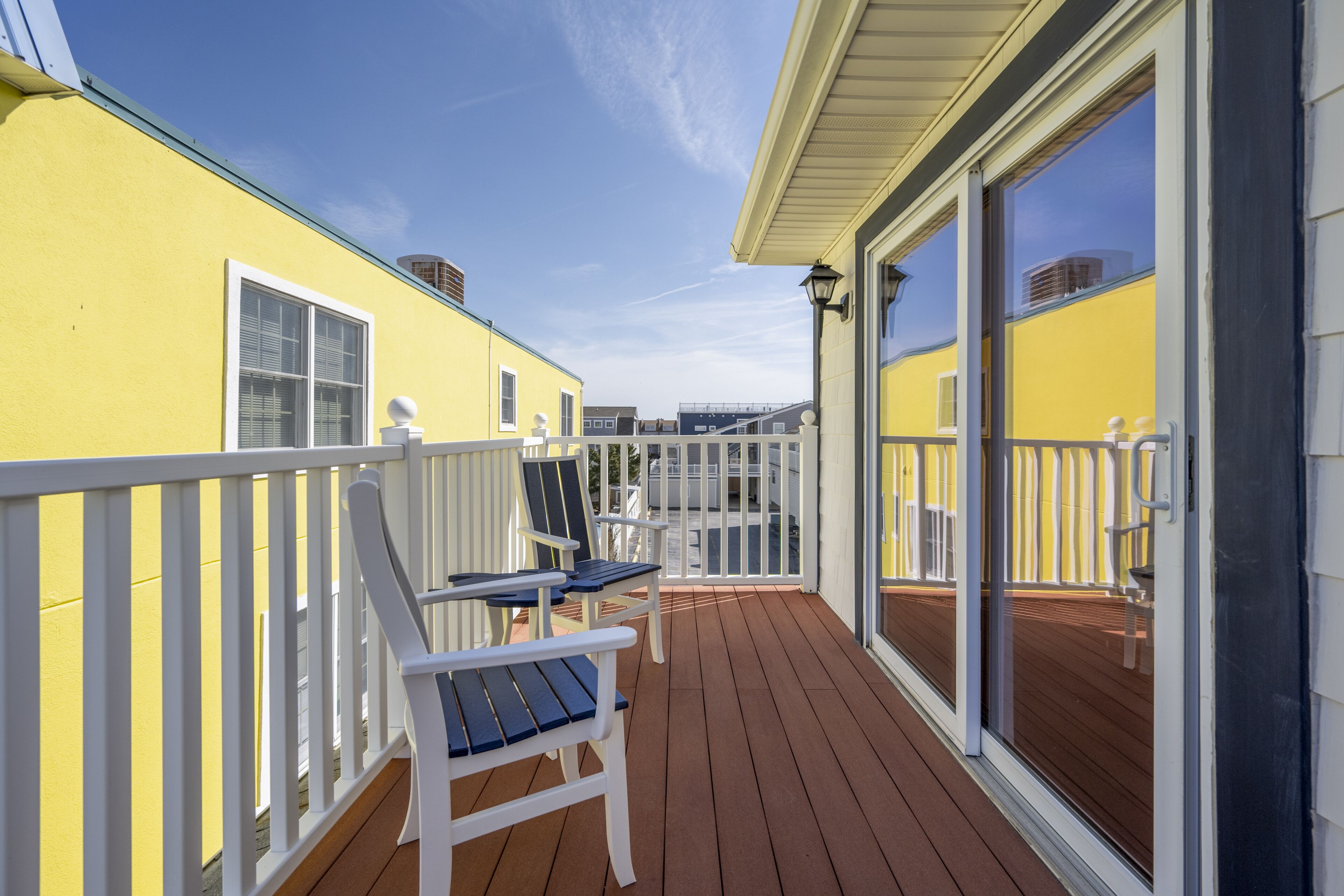 Een houten terras met witte relingen, een geel gebouw en een glazen deur die uitkomt op een balkon met twee stoelen.