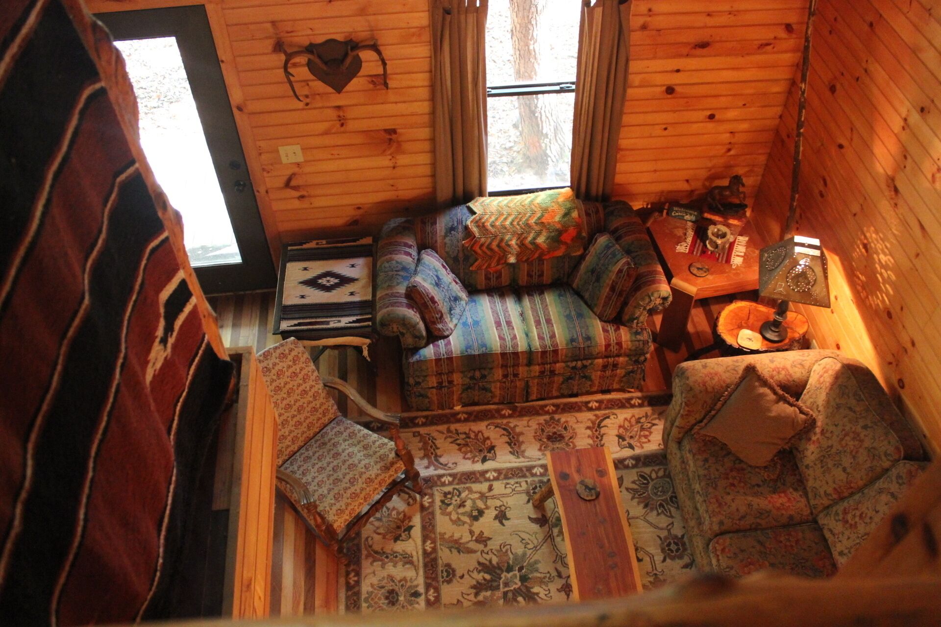A cozy cabin interior with a wooden floor, a sofa set, a small table with a lamp, and a window with curtains.