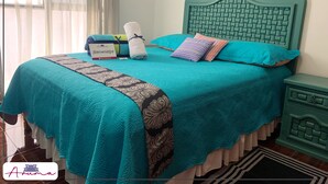 Un letto ben rifatto con copriletto color acquamarina, due cuscini e un asciugamano piegato. Un cartello di benvenuto è appoggiato sul letto.