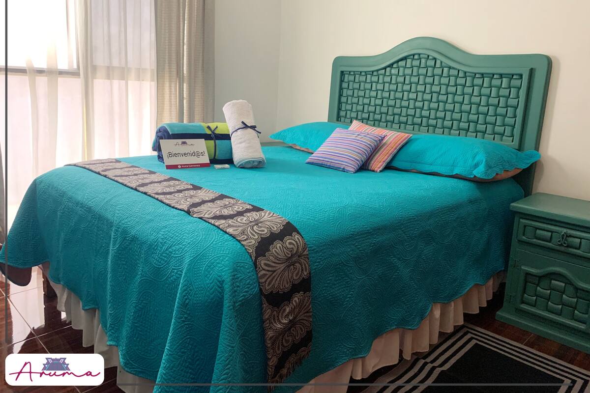 A neatly made bed with a teal bedspread, two pillows, and a folded towel. A welcome sign is placed on the bed.