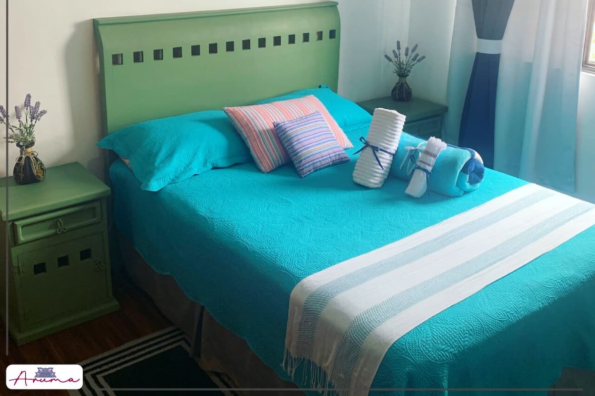 A bed with a teal bedspread and pillows, a white throw blanket, and a green nightstand with a small plant.