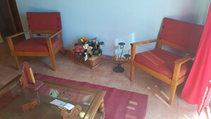 Habitació amb dues cadires de fusta amb coixinets vermells, una cistella de vimet amb flors, una taula petita amb superfície de vidre i una cortina vermella.