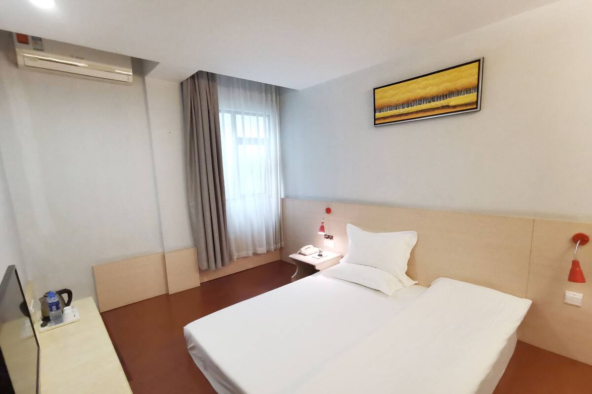 Une chambre d’hôtel avec un lit, une table de chevet, une lampe, un tableau et une fenêtre avec des rideaux.