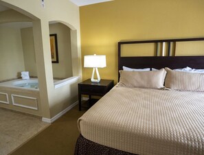Habitación de hotel con una cama grande, una mesita de noche con lámpara, un baño con bañera y un espejo.