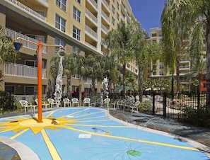 Un área de piscina con un diseño en forma de sol, rodeada de sillones de descanso y palmeras, con un edificio de varios pisos al fondo.