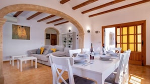 Un coin repas avec une table blanche dressée pour quatre personnes, entourée de chaises blanches. On aperçoit au fond un canapé avec des coussins, un tableau au mur et une porte en arc donnant sur une autre pièce.