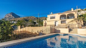 Une villa de style méditerranéen avec une piscine, une montagne en arrière-plan et un ciel bleu dégagé.
