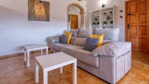 Un salon comprenant un canapé, deux petites tables d’appoint blanches, un tableau, une porte en bois et une étagère ornée d’objets décoratifs.